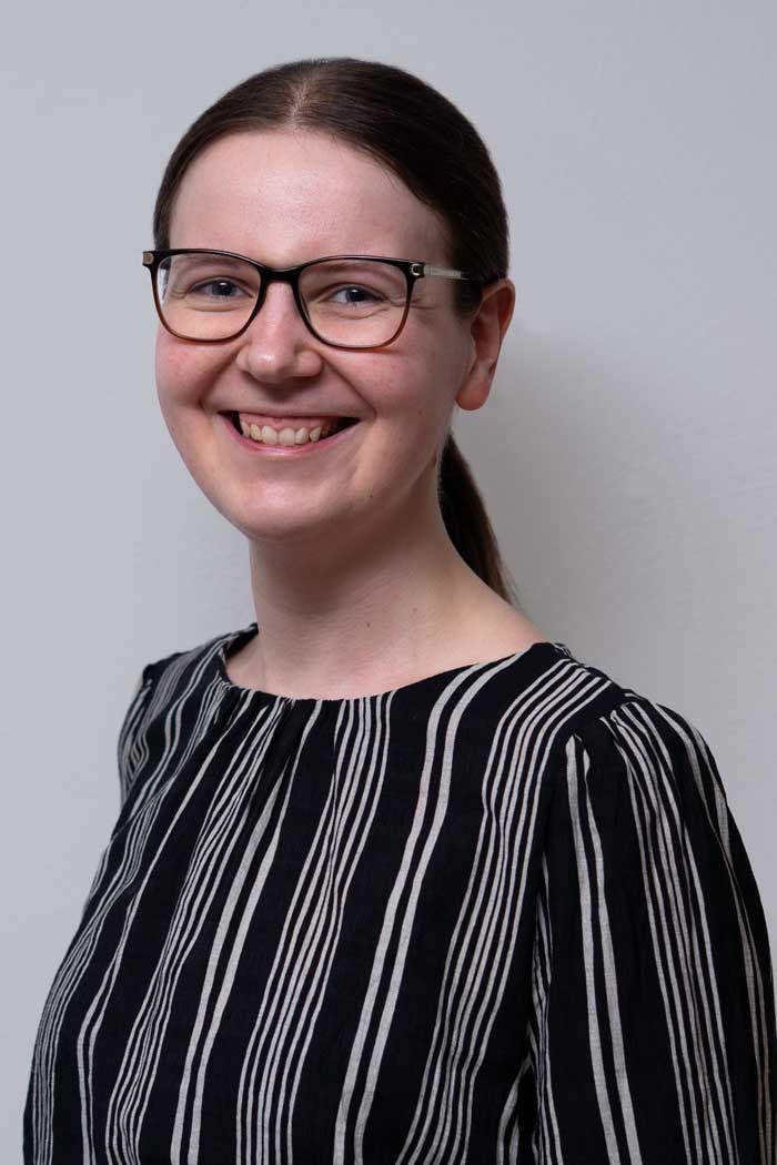 Portrait von Veronika Obermaier. Die langen braunen Haare sind zu einem Pferdeschwanz gebunden. Veronika lacht freundlich in die Kamera. Sie trägt eine eckige, dunkelbraune Brille.